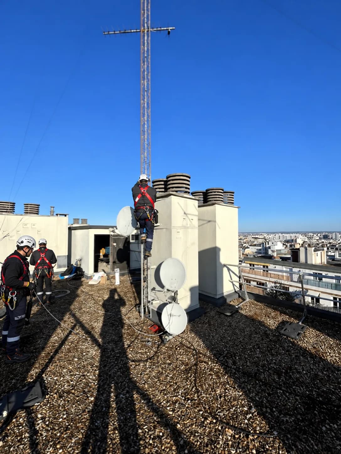 Équipe d'antennistes TELEANTEN en harnais sur un toit de Paris, avec un mât d'antenne râteau et des paraboles